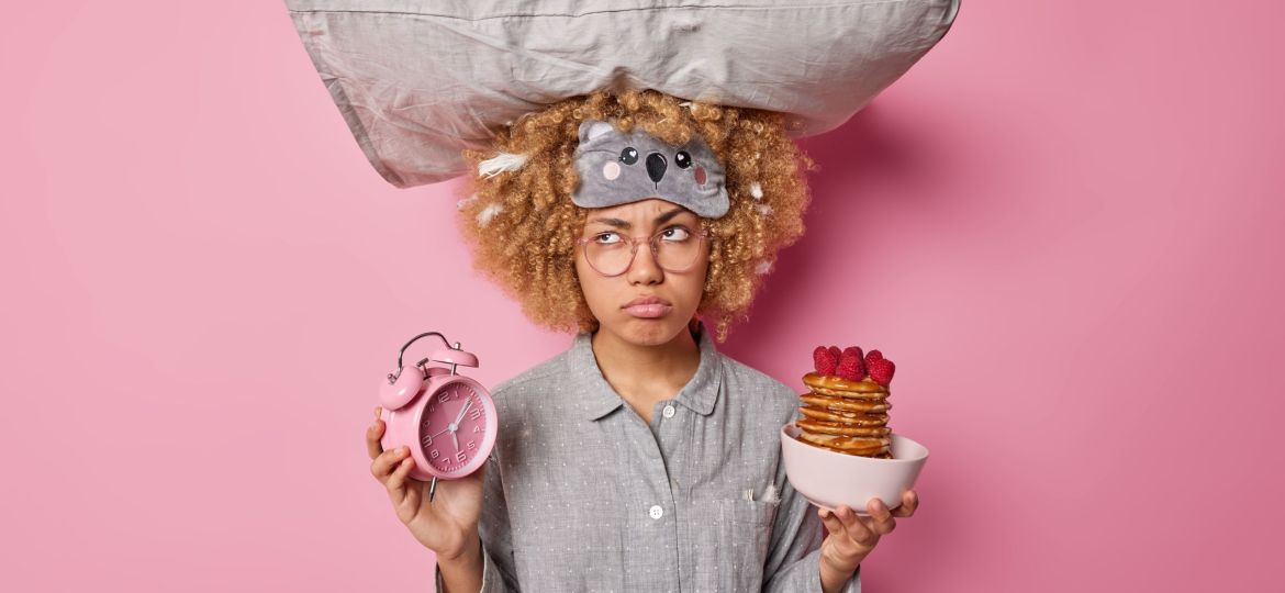 Upset young woman dressed in pajama carries pillow on head holds alarm clock and bowl of pancakes with raspberries has sad pensive expression isolated over pink background. Morning time concept